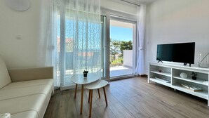 Ett modernt vardagsrum med en vit soffa, ett litet runt bord, en tv på ett vitt tv-bord och en skjutdörr i glas som leder ut till en balkong.
