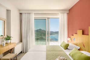 A hotel room with a bed, a desk, a chair, a plant, and a view of a beach and mountains.