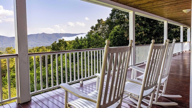 Eine überdachte Veranda mit weißen Schaukelstühlen und Blick auf grüne Hügel und Berge.