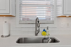 A kitchen with a stainless steel sink, white cabinets, a window with blinds, and a roll of paper towels.