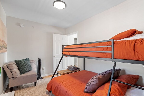 Una cama litera con ropa de cama naranja, un sofá con un cojín verde y una mesita con toallas dobladas.