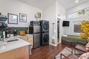 A compact kitchen with a microwave, refrigerator, and washing machine. There is a dining area with a table and chairs, and a bed with a green bedspread.