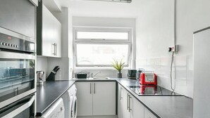 A modern kitchen with stainless steel appliances, a built-in oven, a washing machine, and a window providing natural light. - The Elden - No.14 (London)