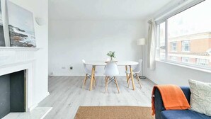 A modern living room with a fireplace, a dining table with chairs, a sofa with a throw blanket, and a large window with a view of buildings. - The Elden - No.14 (London)