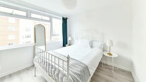 A bedroom with a large window, a bed with white bedding, a bedside table with a lamp, and a decorative mirror. - The Elden - No.14 (London)