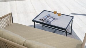 Ein Balkon mit Glasgeländer, einer Couch, einem Tisch mit einer Zeitschrift und einem Getränk.