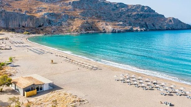 Ein Strand mit einem Berg im Hintergrund, ein kleines Gebäude mit Strohdach und ein klares, blaues Meer.