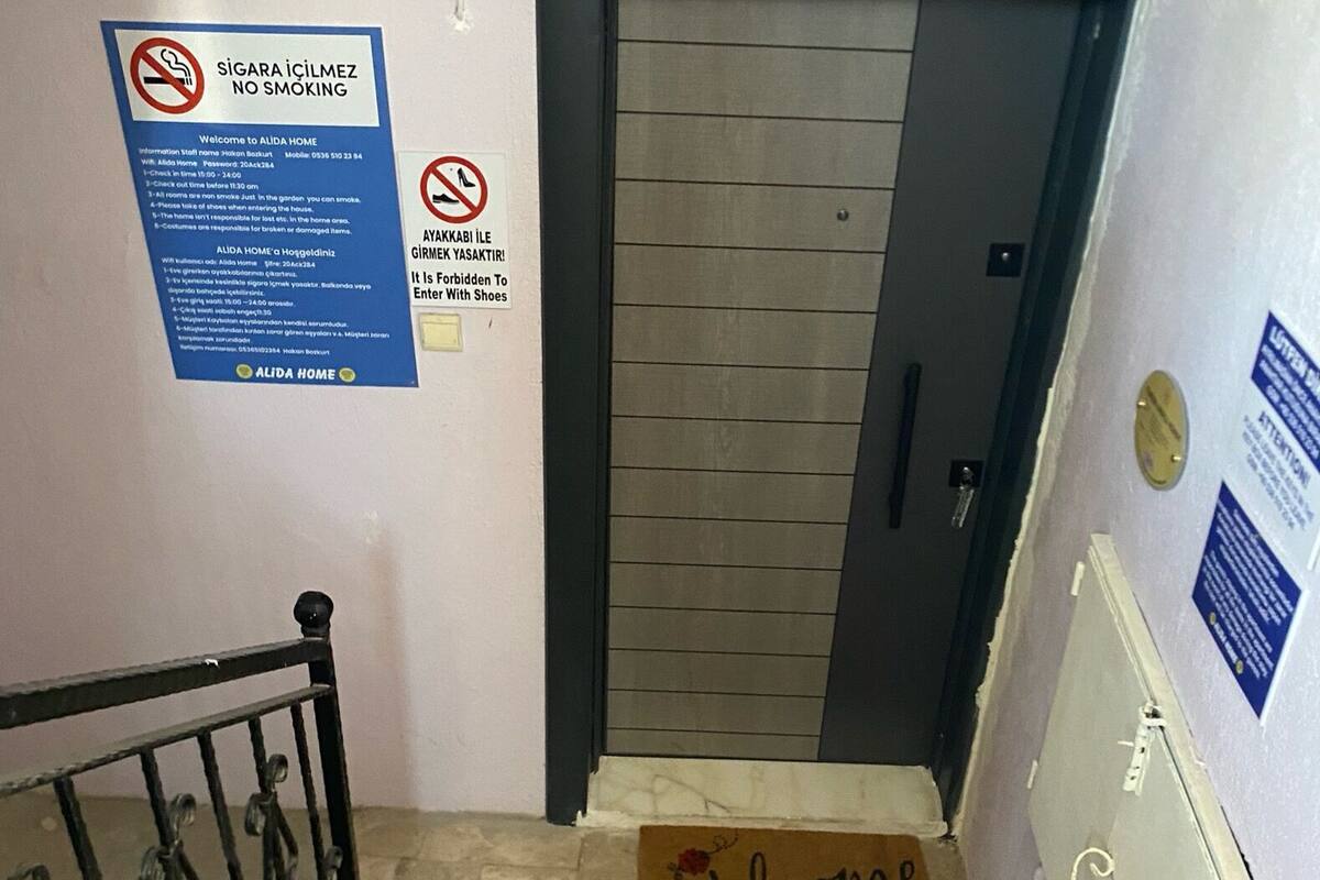 Uma escadaria com um tapete de boas-vindas na base, um aviso de 'Não Fumar' e um aviso de 'Sem Sapatos' na parede.