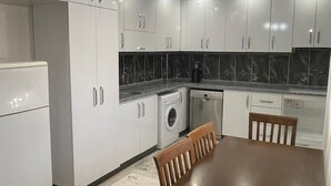 Cocina compacta con muebles blancos, lavadora integrada y comedor con mesa y sillas de madera.