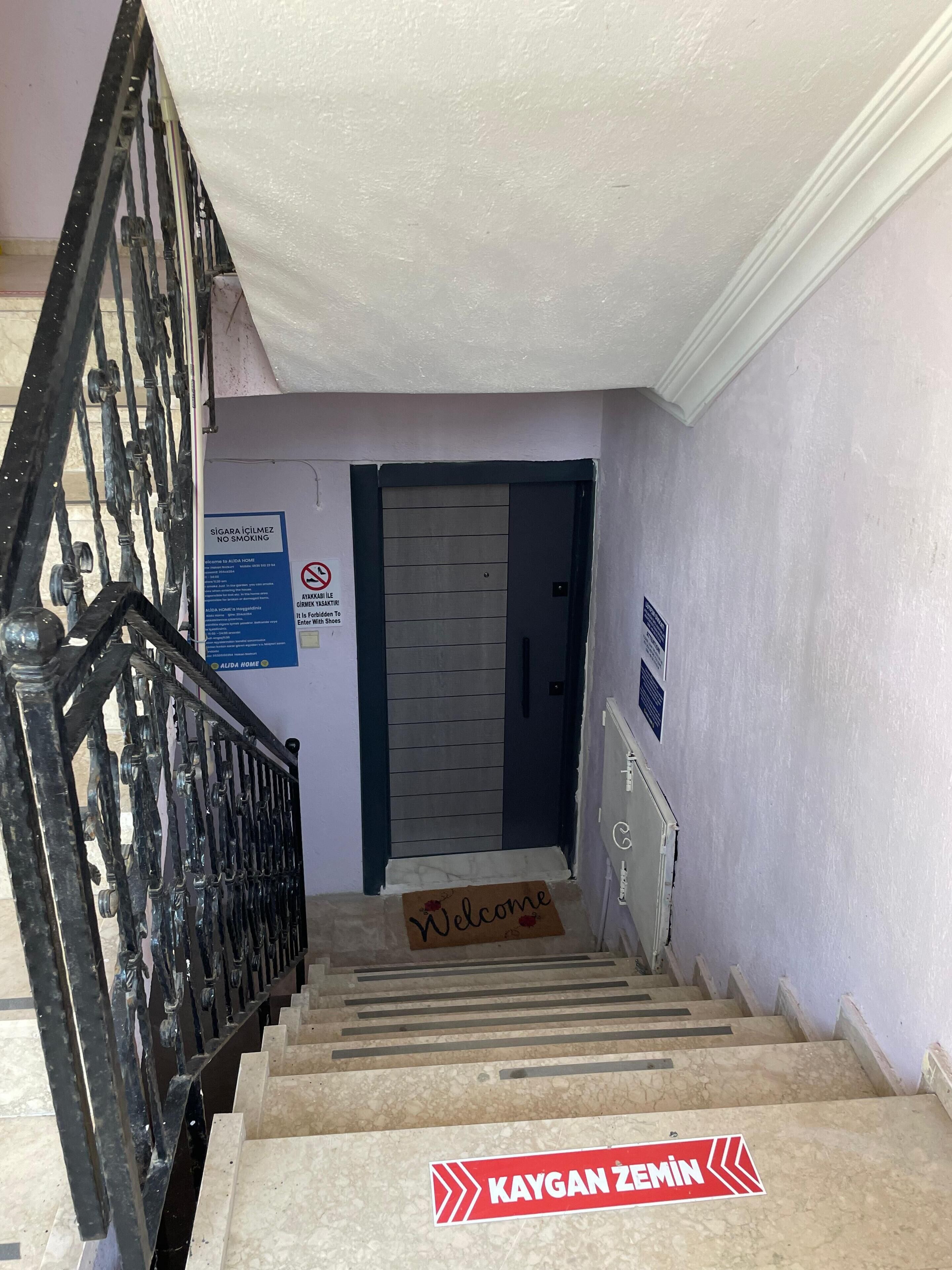 A staircase with a metal railing, a 'Welcome' mat, a 'No Smoking' sign, and a door with a 'Kaygan Zemin' sign.