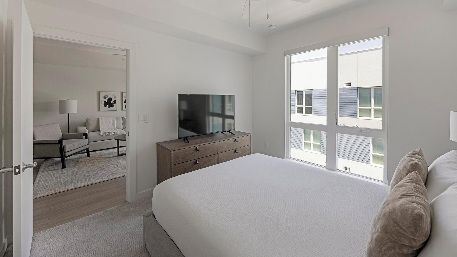 A modern bedroom with a large bed, a TV on a wooden dresser, a sofa, and a view of the outside through a large window.