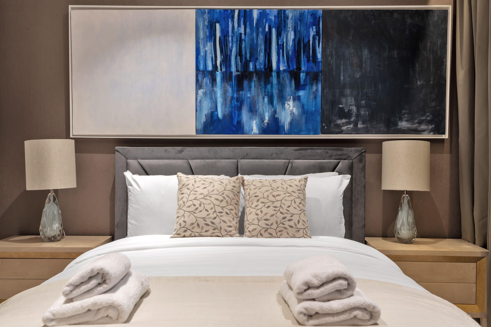 A hotel room with a large bed, two nightstands, a grey upholstered headboard, a large abstract painting, and two lamps.