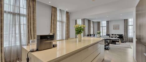 A modern kitchen with a central island, large windows with curtains, and a dining area with a sofa and table.