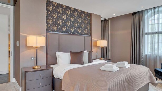 A modern hotel room with a large bed, two bedside tables with lamps, a dark headboard, and a large window with curtains. - Stylish Apartment with AC in Baker St (Greater London)