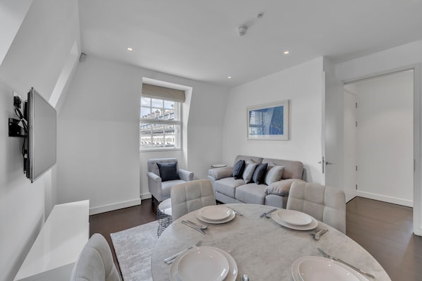 A modern living room with a dining area, a sofa set, a flat-screen TV mounted on the wall, and a large window offering a city view.