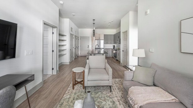A modern living room with a sofa, a coffee table, and a TV. There is a kitchen area with cabinets and a refrigerator. - Landing | Stylish 2BD, Pool, Gym (Nashville)