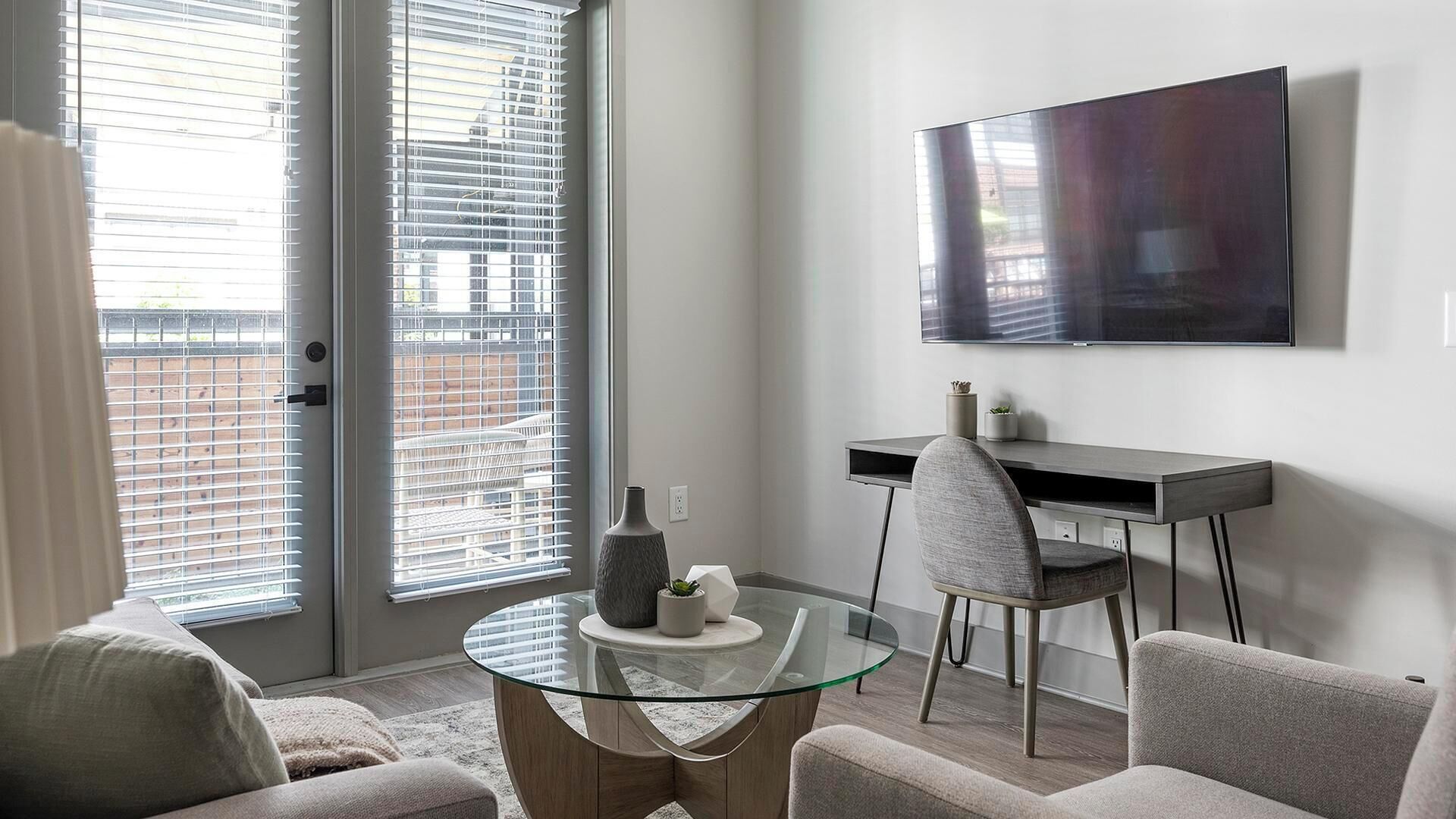 A modern living room with a glass coffee table, a grey sofa, a grey armchair, a flat-screen TV mounted on the wall, and a sliding glass door with blinds.