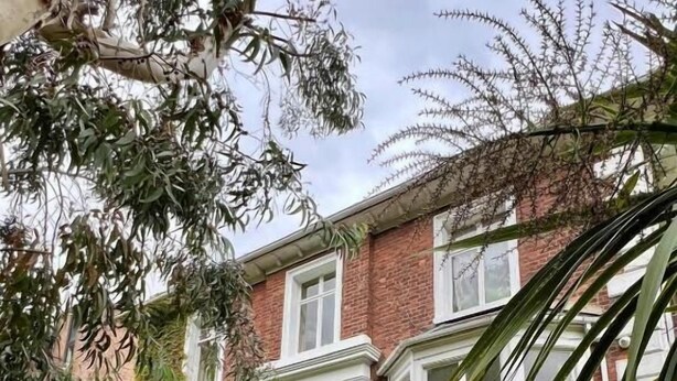 Una casa de dos pisos con un porche delantero, una escalera que conduce a la entrada y un jardín con diversas plantas.