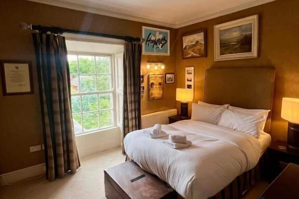A hotel room with a large bed, a bench at the foot of the bed, a window with curtains, a wall with framed pictures, and a sign above the bed.