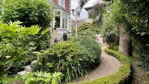 Un jardín residencial con un sendero curvo, follaje denso y una casa de ladrillo con marco de ventana blanco.