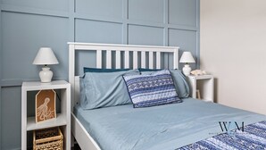Een slaapkamer met een wit bed, blauw beddengoed en een witte nachtkastje met een lamp.