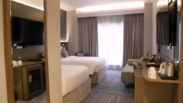 Habitación de hotel con dos camas, un escritorio con cafetera, televisión y un ventanal grande con cortinas.