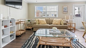 A living room with a sofa, glass coffee table, wicker side tables, a TV mounted on the wall, and a window with blinds. - Charming 2nd Floor Bayside Condo, Steps to Pool & Beach! (Ocean City)