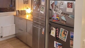 A modern kitchen with a stainless steel refrigerator covered in photos and magnets, wooden cabinets, and a window with blinds. - Buttes Chaumont top floor loft: unobstructed view of Paris! (Paris)