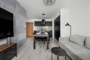 En moderne stue med et fladskærms TV, en grå sofa, et spiseområde med bord og stole, og et køkken i baggrunden.