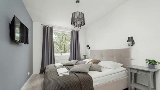 A modern bedroom with a bed, a TV, a chandelier, a nightstand, and a window with curtains. - Stylish Apartment with 2 Bedrooms | Balcony and Parking (Wroclaw)