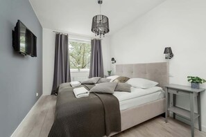 Ein modernes Schlafzimmer mit einem Bett, einem Fernseher, einer Chandelieber, einem Nachttisch und einem Fenster mit Vorhängen.