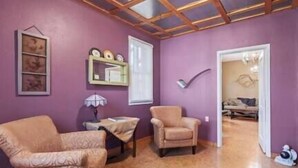 Una habitación con techo de madera, pared morada, un sillón estampado, una mesita con lámpara y una puerta que da a otra habitación.