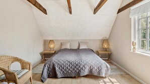 A bedroom with a sloped ceiling, exposed wooden beams, a bed with a quilted duvet, two bedside lamps, a wicker chair, and a window with a view.