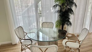Una mesa redonda de vidrio rodeada de sillas de mimbre en una habitación con suelo de madera y cortinas traslúcidas.