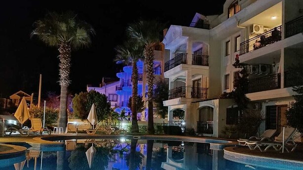 A nighttime view of a swimming pool with lounge chairs and palm trees, surrounded by multi-story buildings with balconies.