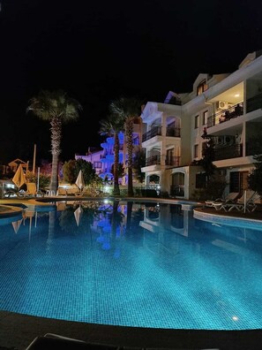 A nighttime view of a swimming pool with lounge chairs and palm trees, surrounded by multi-story buildings with balconies.