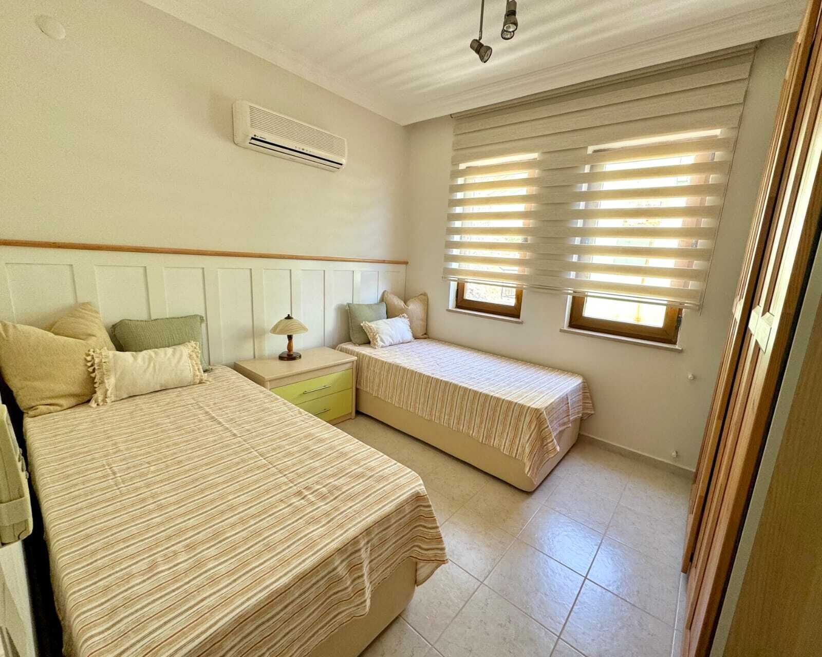 A bedroom with two beds, a nightstand, a lamp, and an air conditioning unit.