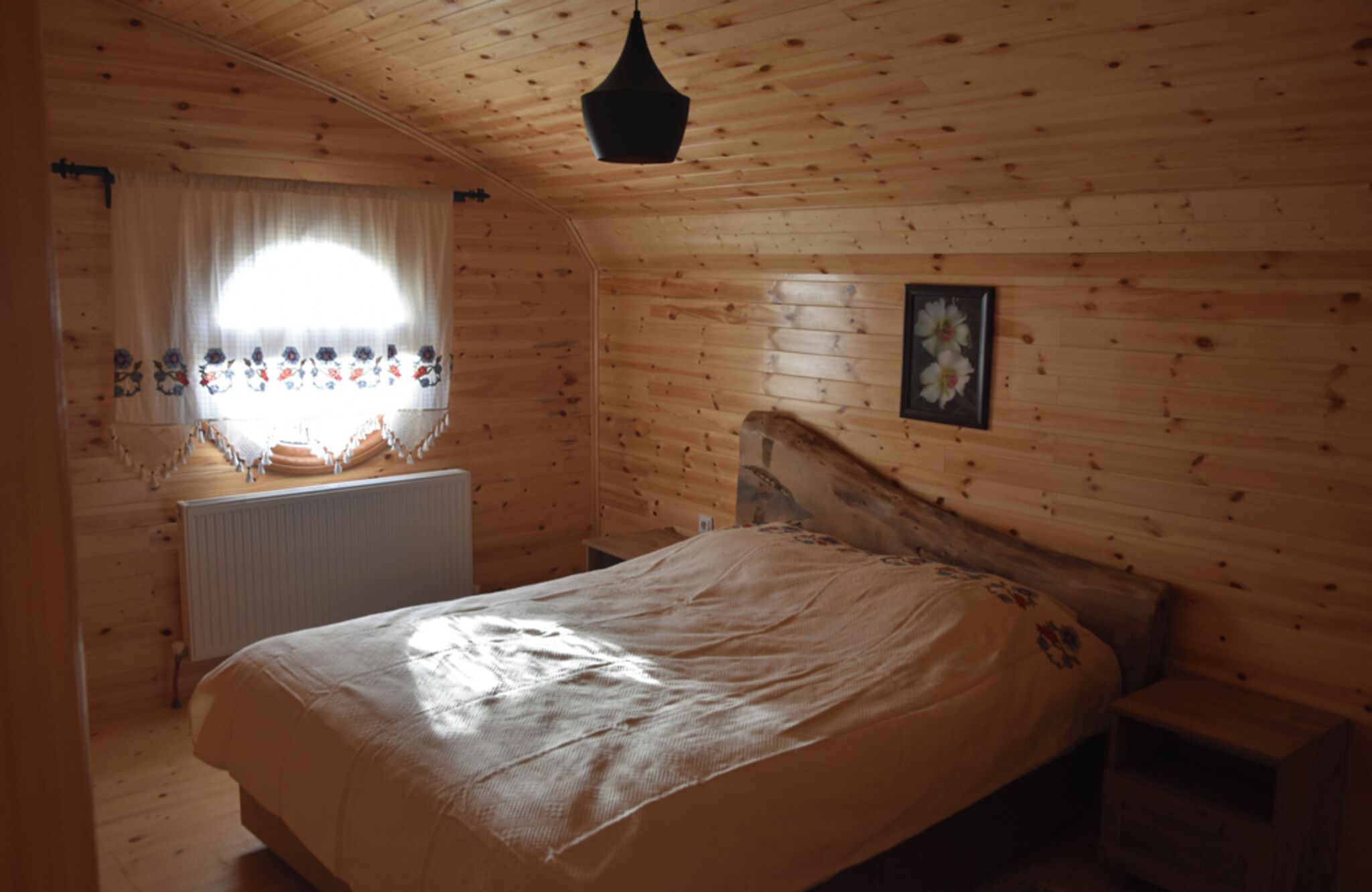 A wooden cabin bedroom with a bed, a window with curtains, and a hanging light fixture.