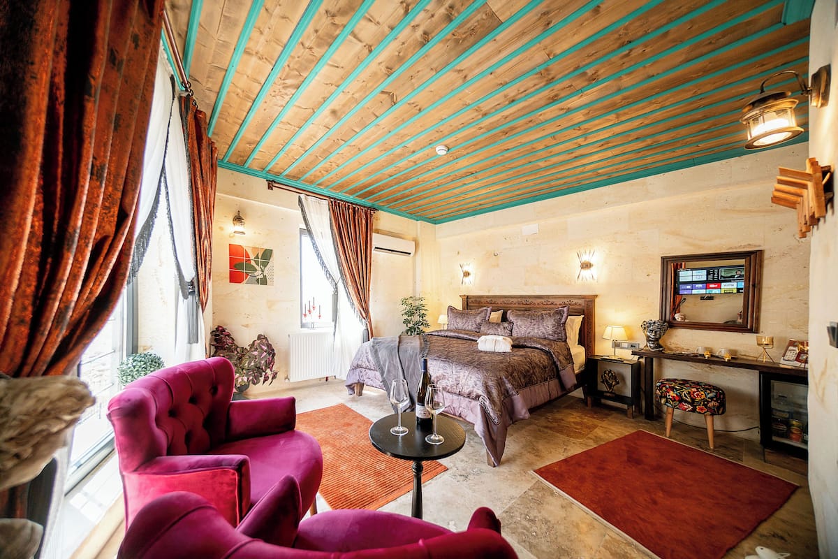 A hotel room with a wooden ceiling, a bed with a headboard, a nightstand, a table with a bottle and glasses, and a chair.
