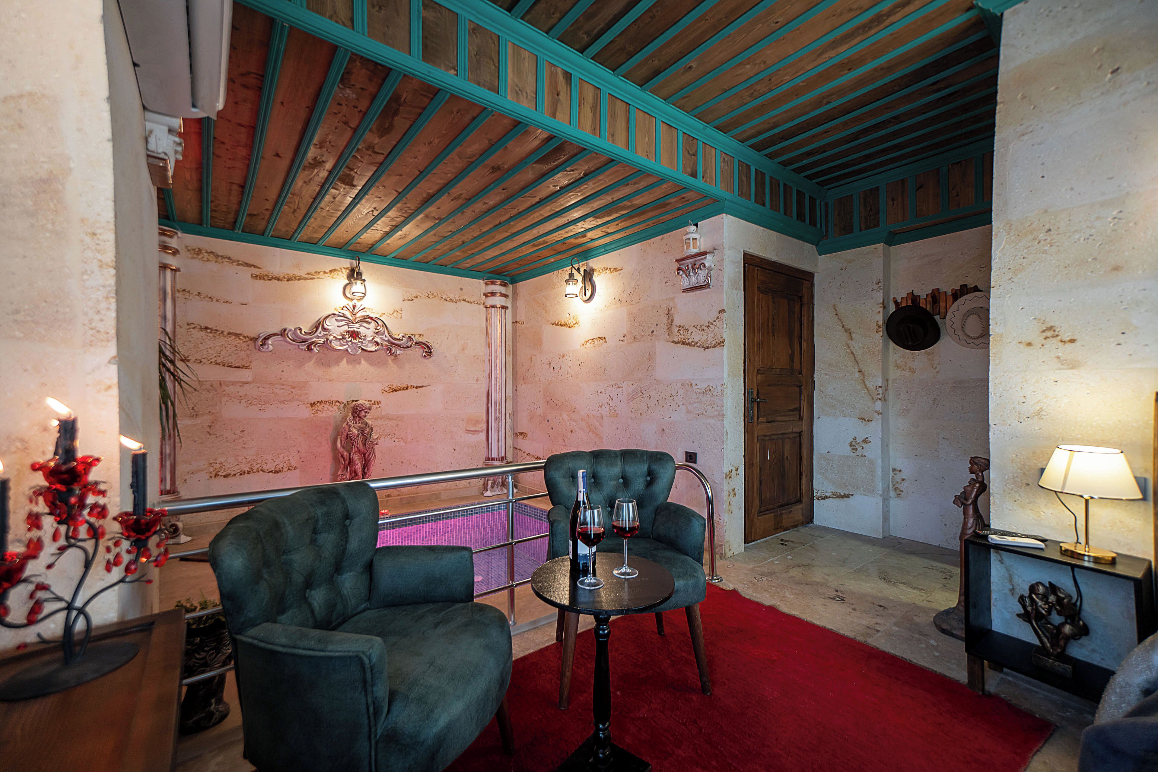 Une pièce avec des poutres en bois, une moquette rouge, deux fauteuils rembourrés, une petite table avec une bouteille et des verres, et une lampe.