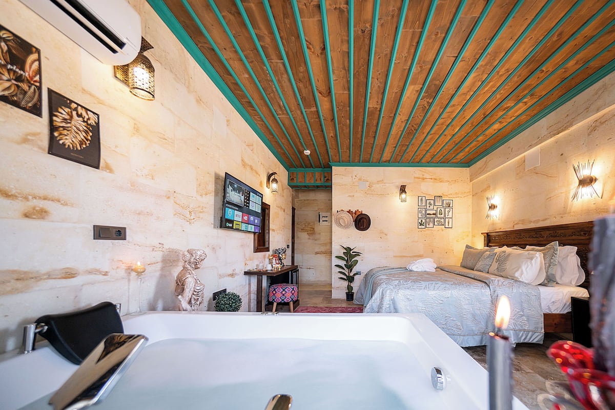A hotel room with a large bathtub, a bed with a patterned bedspread, a wooden ceiling with teal accents, and wall-mounted sconces.