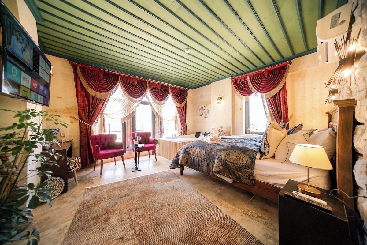 A bedroom with a bed, a nightstand, a lamp, a chair, a table, and a large window with curtains.