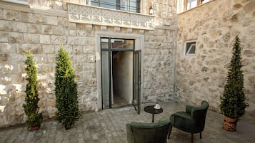 Un patio con muros de piedra, una puerta que da al interior y dos sillas verdes alrededor de una pequeña mesa.
