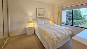 Un dormitorio con cama, dos mesitas de noche con lámparas, un ventilador de techo, una ventana con cortinas y un cuadro en la pared.