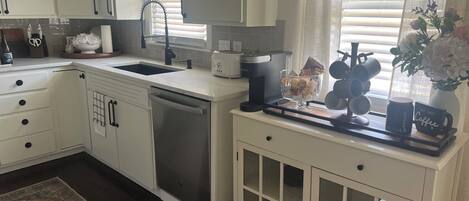 A modern kitchen with white cabinetry, a dishwasher, a coffee maker, and a window with blinds.