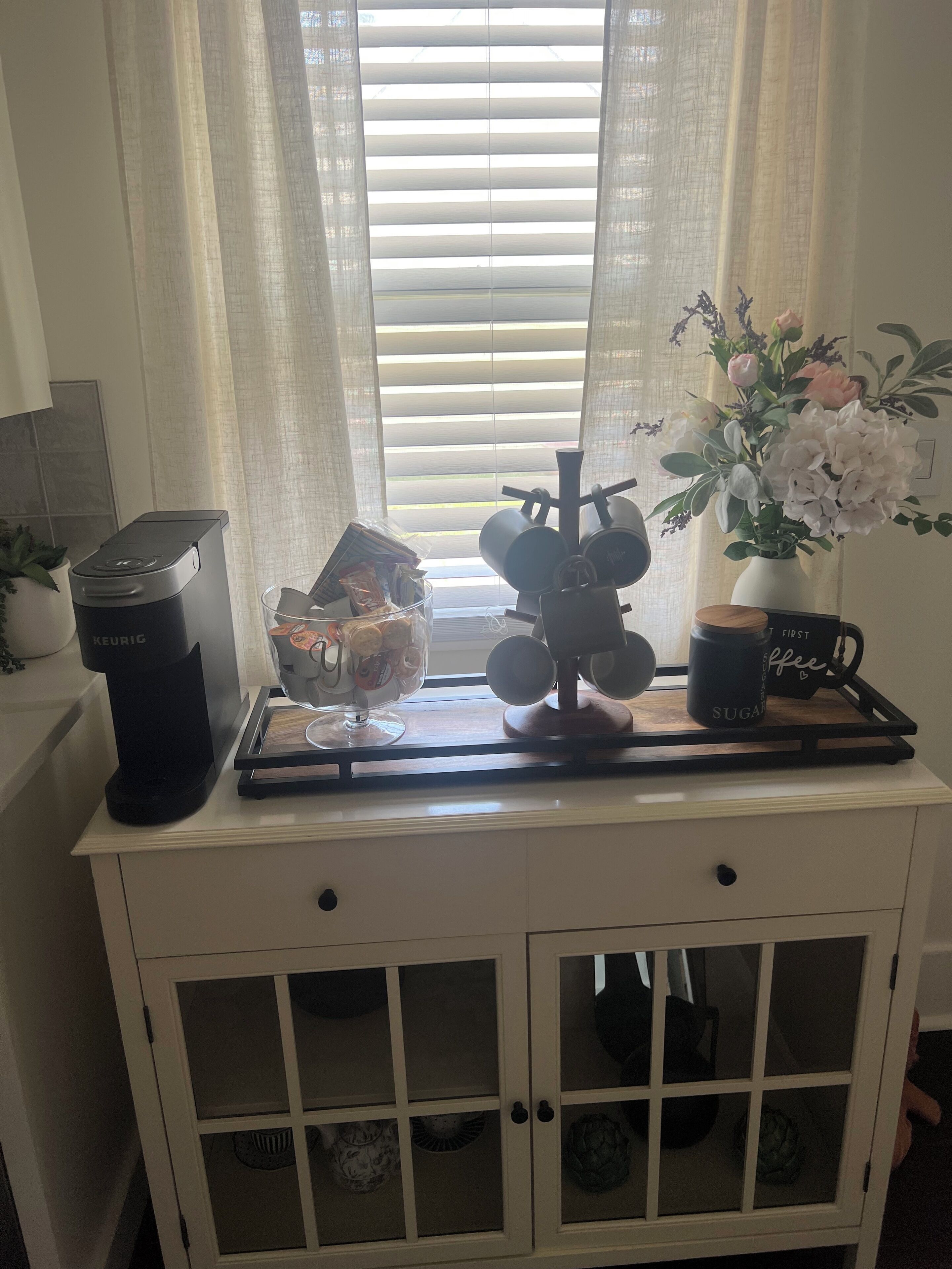 A kitchen counter with a Keurig coffee maker, a glass container with snacks, a coffee machine, a mug, and a vase with flowers.