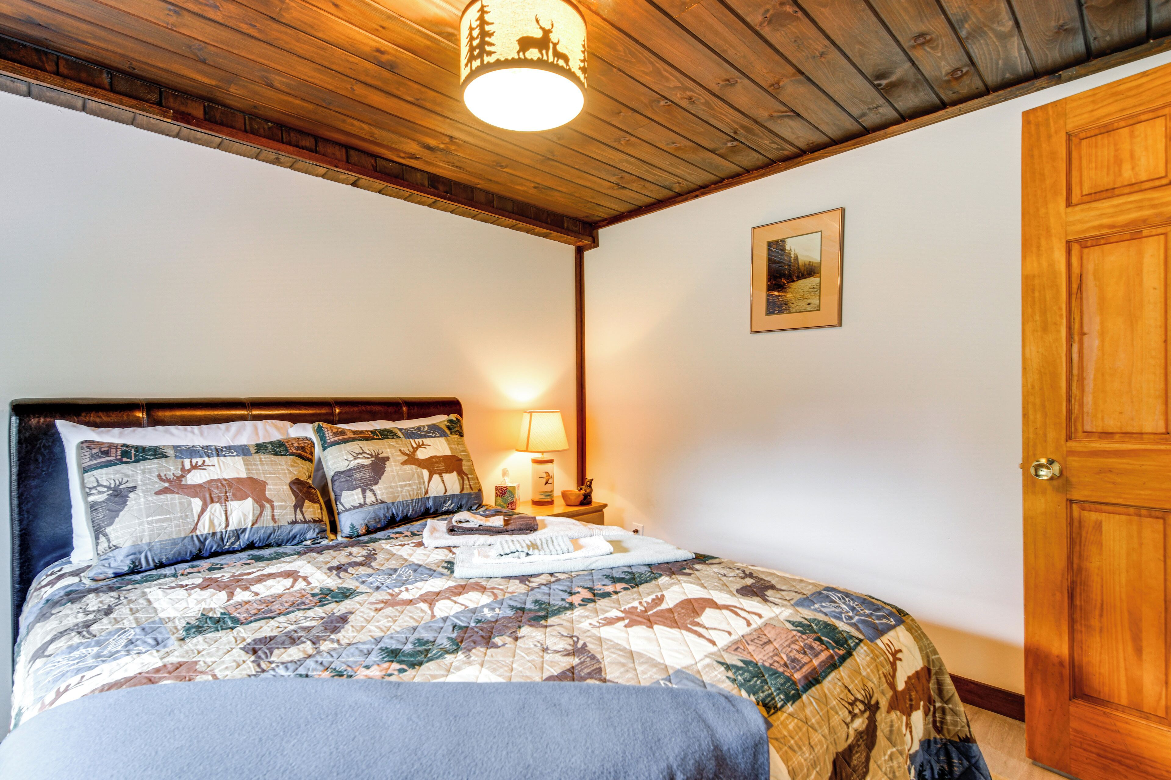 A bedroom with a wooden ceiling, a bed with a quilt featuring deer, a nightstand with a lamp, and a door.