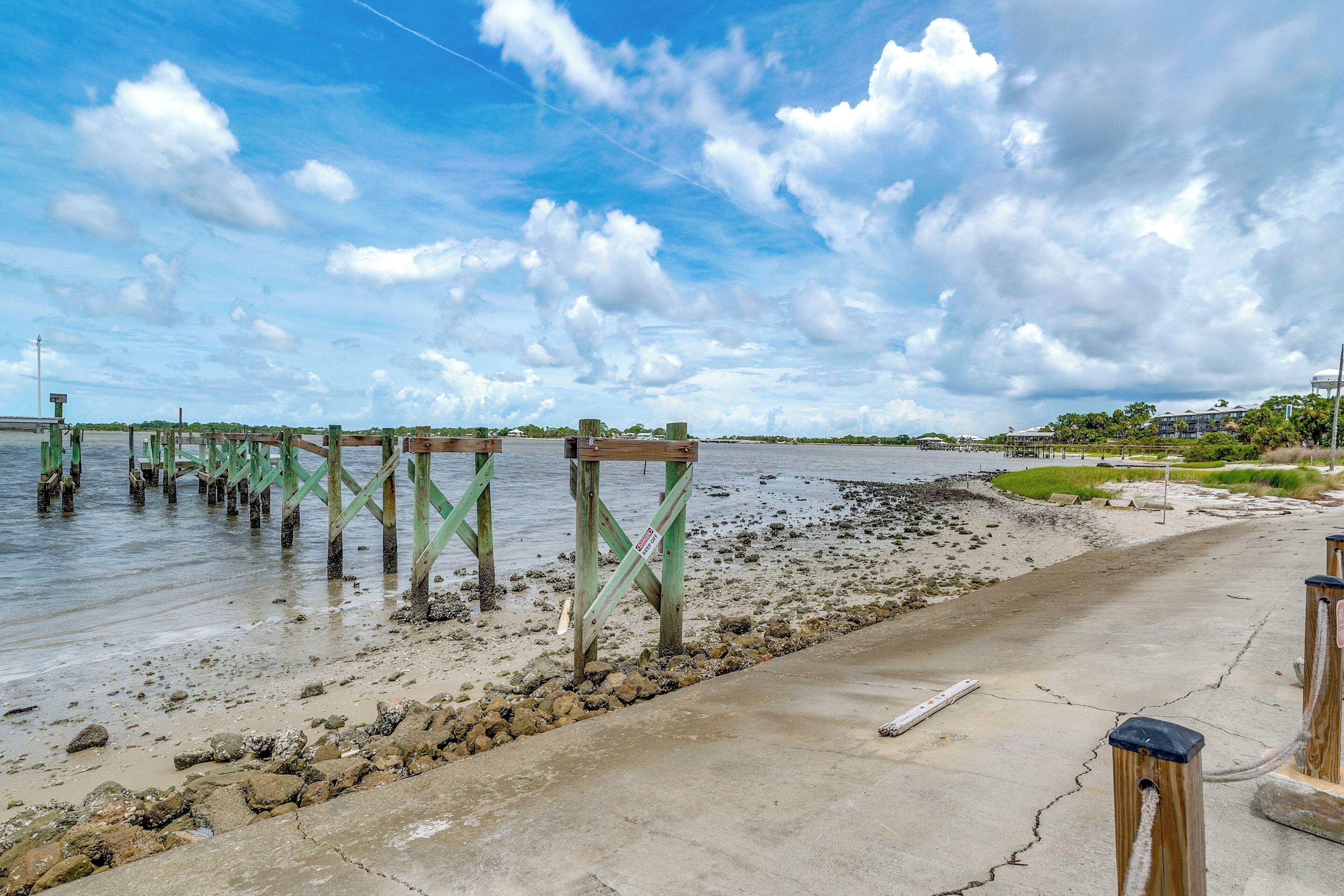 A coastal walkway with wooden pilings extending into the water, a concrete path, and a grassy area with buildings in the background.