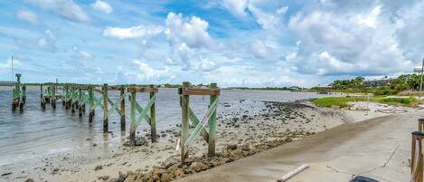A coastal walkway with wooden pilings extending into the water, a concrete path, and a grassy area with buildings in the background.