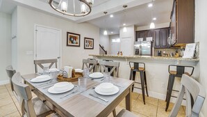 A dining area with a wooden table set for four, a chandelier, and a kitchen in the background.